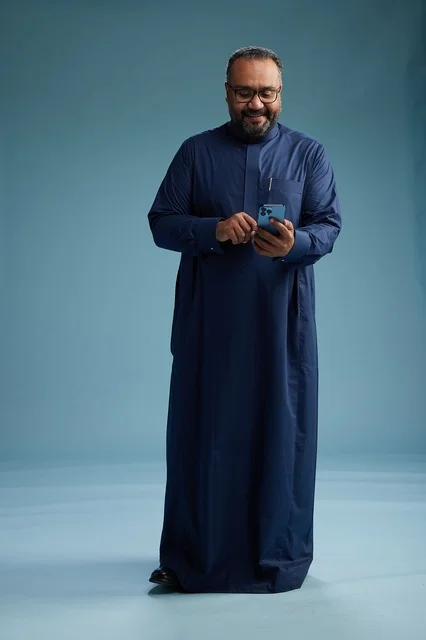 A portrait of a Saudi man wearing a navy thobe using a smartphone, communicating via mobile, full body, blue background.