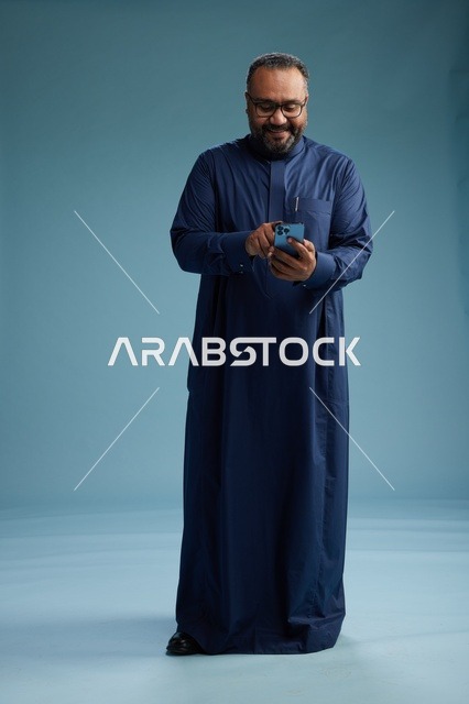 بورتريه لرجل سعودي يرتدي ثوب كحلي يستخدم الهاتف الذكي, التواصل عبر الجوال, الجسم بالكامل, خلفيه زرقاء