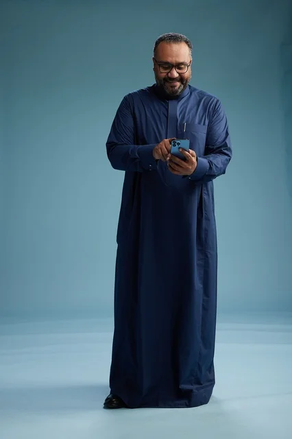 A portrait of a Saudi man wearing a navy thobe using a smartphone, communicating via mobile, full body, blue background.