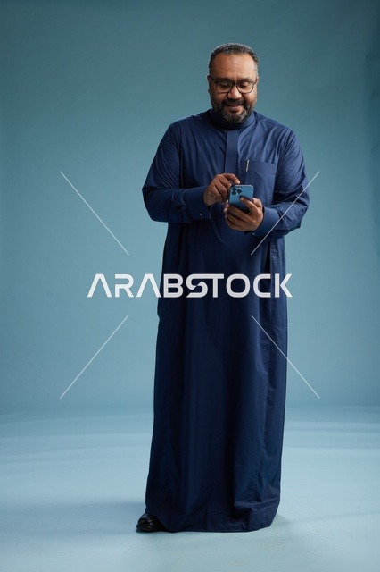 A portrait of a Saudi man wearing a navy thobe using a smartphone, communicating via mobile, full body, blue background.
