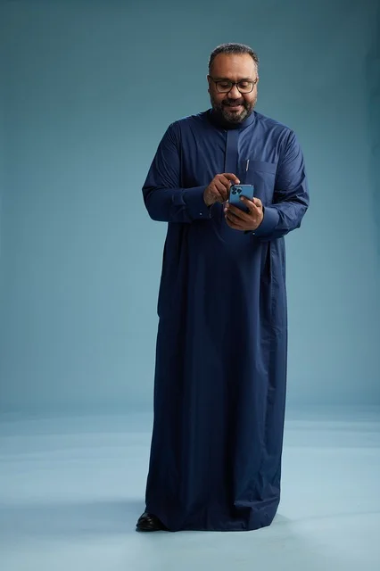 A portrait of a Saudi man wearing a navy thobe using a smartphone, communicating via mobile, full body, blue background.