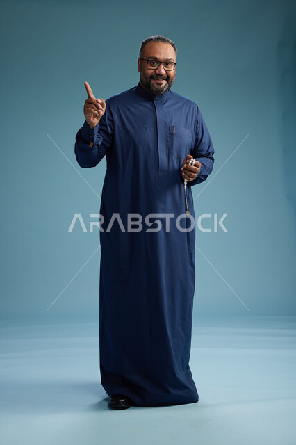 بورتريه لرجل سعودي يقف مرتدياً ثوب كحلي يقوم بايماءات وحركات متنوعه, الوقوف بكامل الجسم, خلفيه زرقاء
