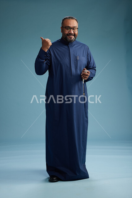 بورتريه لرجل سعودي يقف مرتدياً ثوب كحلي يقوم بايماءات وحركات متنوعه, الوقوف بكامل الجسم, خلفيه زرقاء