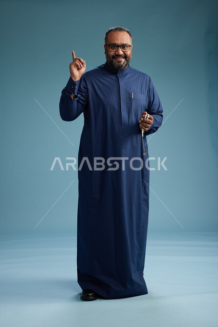 بورتريه لرجل سعودي يقف مرتدياً ثوب كحلي يقوم بايماءات وحركات متنوعه, الوقوف بكامل الجسم, خلفيه زرقاء