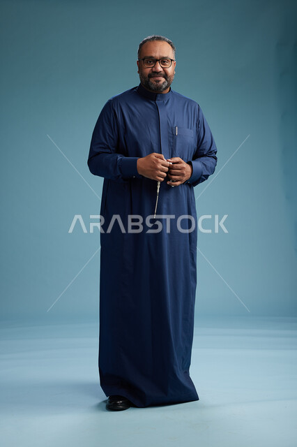 A portrait of a Saudi man standing wearing a navy blue thobe, making various gestures and movements, standing full body, with a blue background.