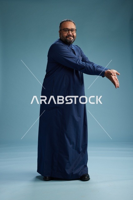 بورتريه لرجل سعودي يقف مرتدياً ثوب كحلي ينظر الى الكاميرا, كامل الجسم, خلفيه زرقاء