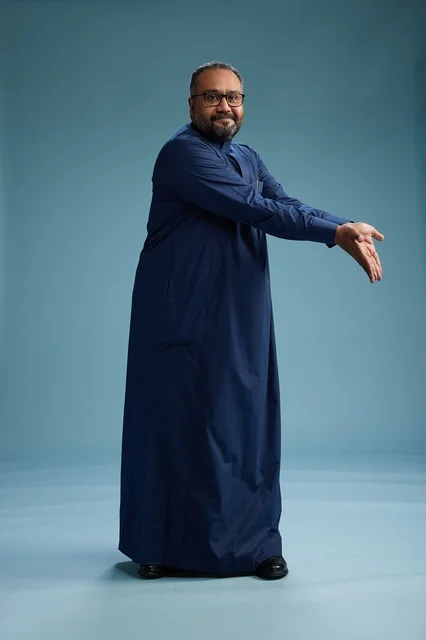 بورتريه لرجل سعودي يقف مرتدياً ثوب كحلي ينظر الى الكاميرا, كامل الجسم, خلفيه زرقاء
