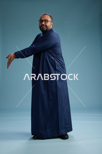 بورتريه لرجل سعودي يقف مرتدياً ثوب كحلي ينظر الى الكاميرا, كامل الجسم, خلفيه زرقاء
