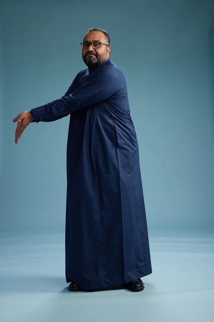 بورتريه لرجل سعودي يقف مرتدياً ثوب كحلي ينظر الى الكاميرا, كامل الجسم, خلفيه زرقاء
