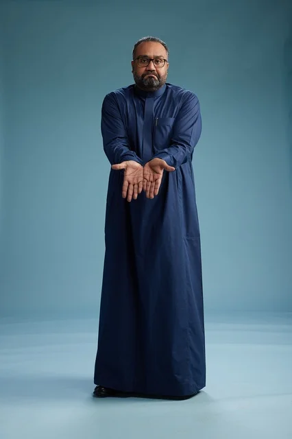 A portrait of a Saudi man standing wearing a navy blue thobe looking at the camera, full body, with a blue background.