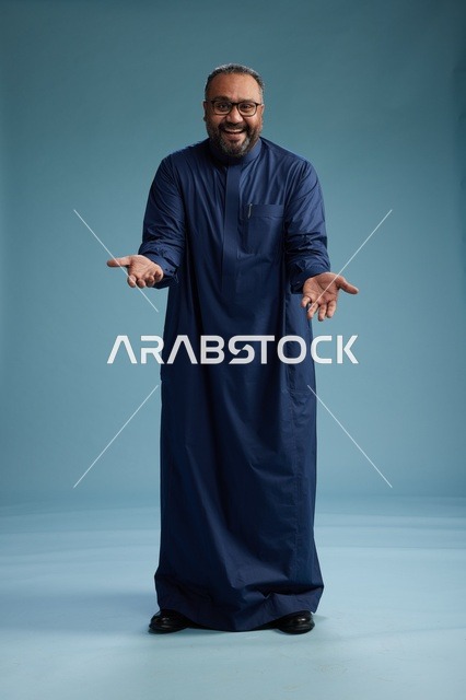 A portrait of a Saudi man standing wearing a navy blue thobe looking at the camera, full body, with a blue background.
