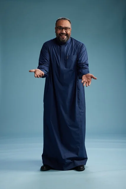 بورتريه لرجل سعودي يقف مرتدياً ثوب كحلي ينظر الى الكاميرا, كامل الجسم, خلفيه زرقاء