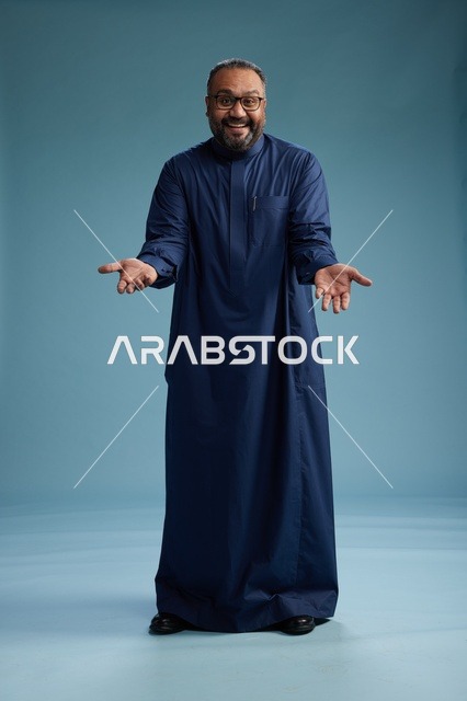 بورتريه لرجل سعودي يقف مرتدياً ثوب كحلي ينظر الى الكاميرا, كامل الجسم, خلفيه زرقاء