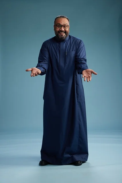 A portrait of a Saudi man standing wearing a navy blue thobe looking at the camera, full body, with a blue background.