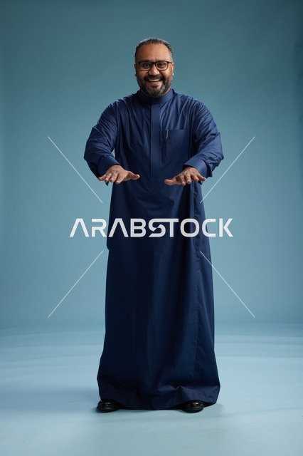بورتريه لرجل سعودي يقف مرتدياً ثوب كحلي ينظر الى الكاميرا, كامل الجسم, خلفيه زرقاء