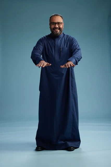 بورتريه لرجل سعودي يقف مرتدياً ثوب كحلي ينظر الى الكاميرا, كامل الجسم, خلفيه زرقاء