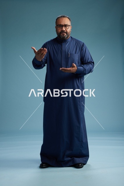بورتريه لرجل سعودي يقف مرتدياً ثوب كحلي ينظر الى الكاميرا, كامل الجسم, خلفيه زرقاء