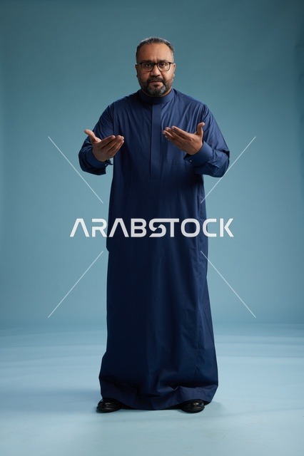 A portrait of a Saudi man standing wearing a navy blue thobe looking at the camera, full body, with a blue background.