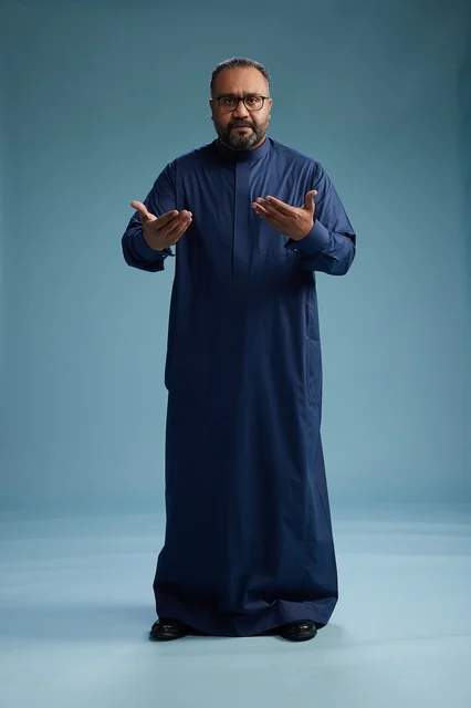 A portrait of a Saudi man standing wearing a navy blue thobe looking at the camera, full body, with a blue background.