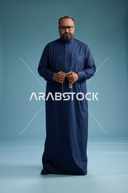بورتريه لرجل سعودي يقف مرتدياً ثوب كحلي ينظر الى الكاميرا, كامل الجسم, خلفيه زرقاء