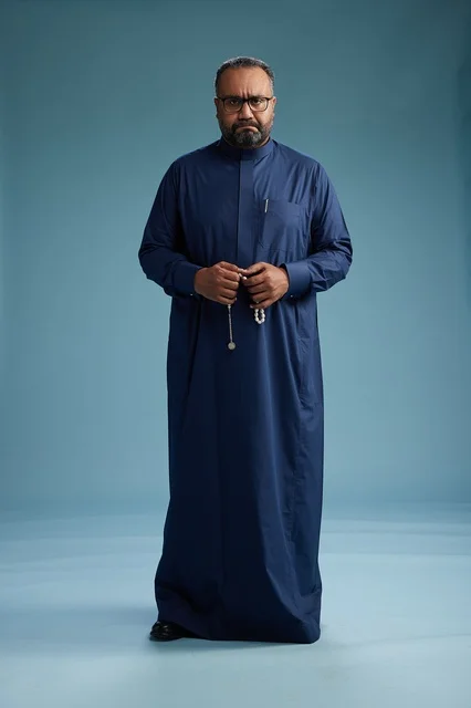 A portrait of a Saudi man standing wearing a navy blue thobe looking at the camera, full body, with a blue background.