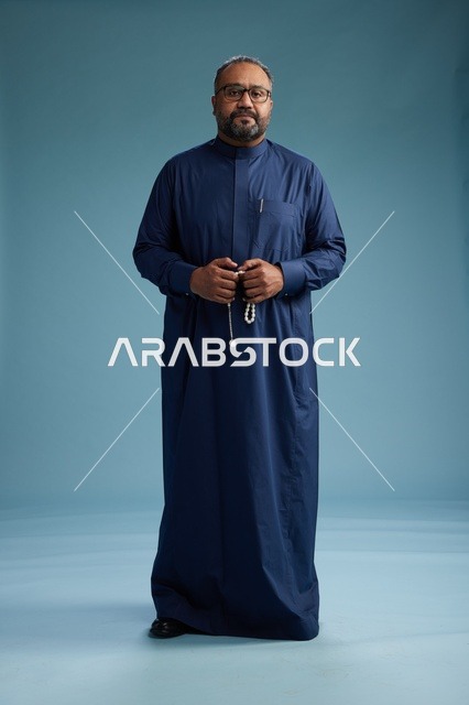 بورتريه لرجل سعودي يقف مرتدياً ثوب كحلي ينظر الى الكاميرا, كامل الجسم, خلفيه زرقاء