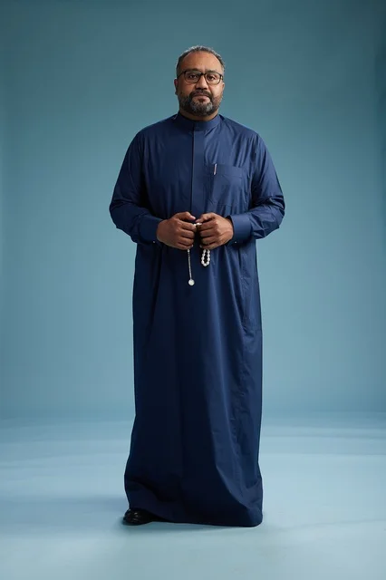 A portrait of a Saudi man standing wearing a navy blue thobe looking at the camera, full body, with a blue background.