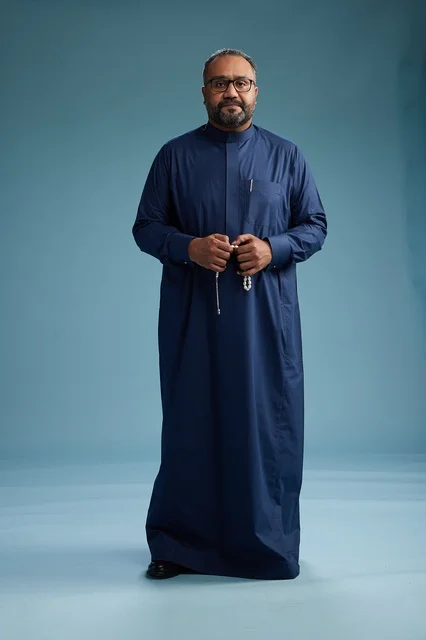 A portrait of a Saudi man standing wearing a navy blue thobe looking at the camera, full body, with a blue background.