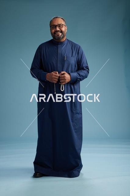 A portrait of a Saudi man standing wearing a navy blue thobe looking at the camera, full body, with a blue background.