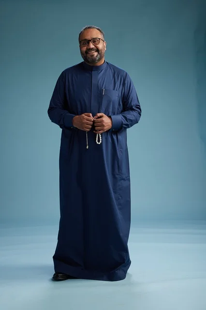 A portrait of a Saudi man standing wearing a navy blue thobe looking at the camera, full body, with a blue background.