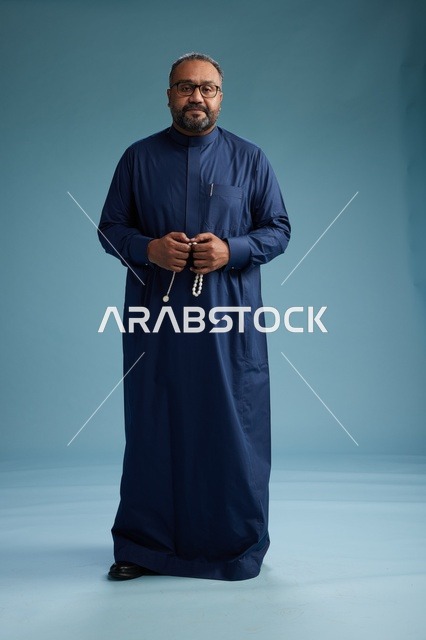 بورتريه لرجل سعودي يقف مرتدياً ثوب كحلي ينظر الى الكاميرا, كامل الجسم, خلفيه زرقاء
