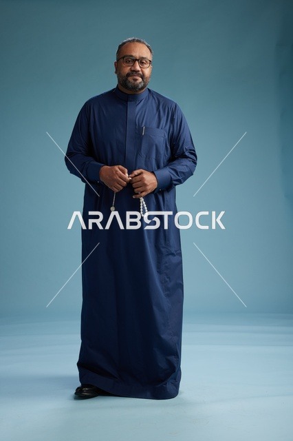 A portrait of a Saudi man standing wearing a navy blue thobe looking at the camera, full body, with a blue background.