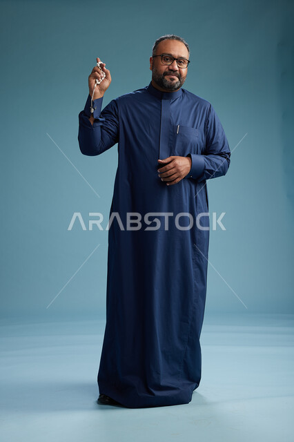 A portrait of a Saudi man standing wearing a navy blue thobe, making various gestures and movements, standing full body, with a blue background.