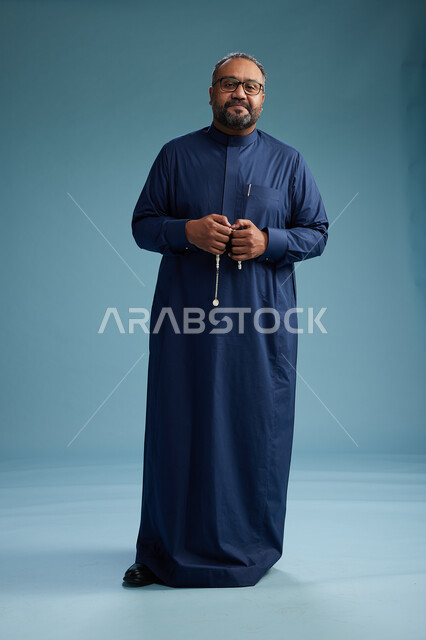 A portrait of a Saudi man standing wearing a navy blue thobe, making various gestures and movements, standing full body, with a blue background.