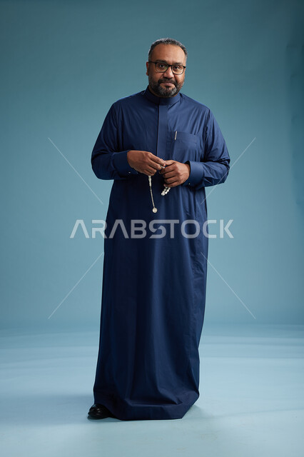 بورتريه لرجل سعودي يقف مرتدياً ثوب كحلي يقوم بايماءات وحركات متنوعه, الوقوف بكامل الجسم, خلفيه زرقاء