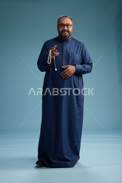 بورتريه لرجل سعودي يقف مرتدياً ثوب كحلي يقوم بايماءات وحركات متنوعه, الوقوف بكامل الجسم, خلفيه زرقاء