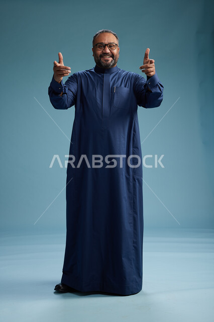 A portrait of a Saudi man standing wearing a navy blue thobe, making various gestures and movements, standing full body, with a blue background.