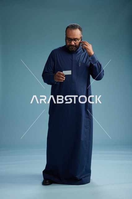 A portrait of a Saudi man wearing a blue thobe looking at a business card while holding a mobile phone, full body, blue background.