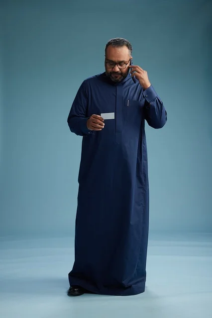 بورتريه لرجل سعودي يرتدي ثوباً أزرق ينظر إلى بطاقة عمل ممسكاً بالهاتف المحمول, الجسم بالطول الكامل, خلفيه زرقاء