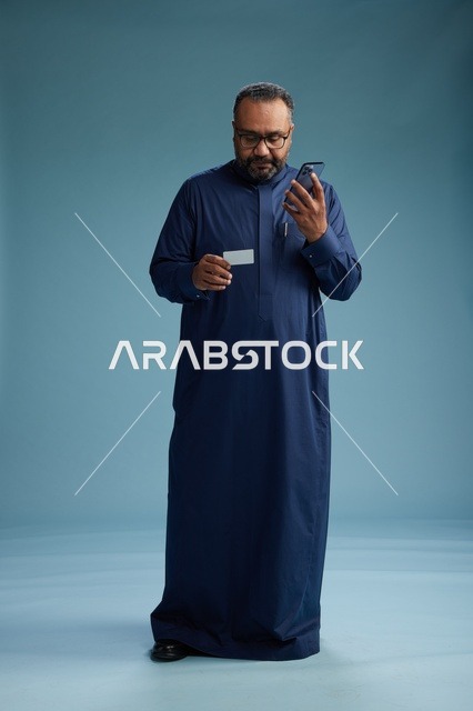 بورتريه لرجل سعودي يرتدي ثوباً أزرق ينظر إلى بطاقة عمل ممسكاً بالهاتف المحمول, الجسم بالطول الكامل, خلفيه زرقاء