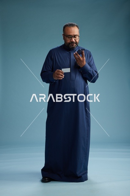 بورتريه لرجل سعودي يرتدي ثوباً أزرق ينظر إلى بطاقة عمل ممسكاً بالهاتف المحمول, الجسم بالطول الكامل, خلفيه زرقاء