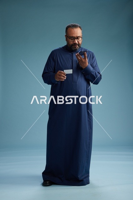بورتريه لرجل سعودي يرتدي ثوباً أزرق ينظر إلى بطاقة عمل ممسكاً بالهاتف المحمول, الجسم بالطول الكامل, خلفيه زرقاء