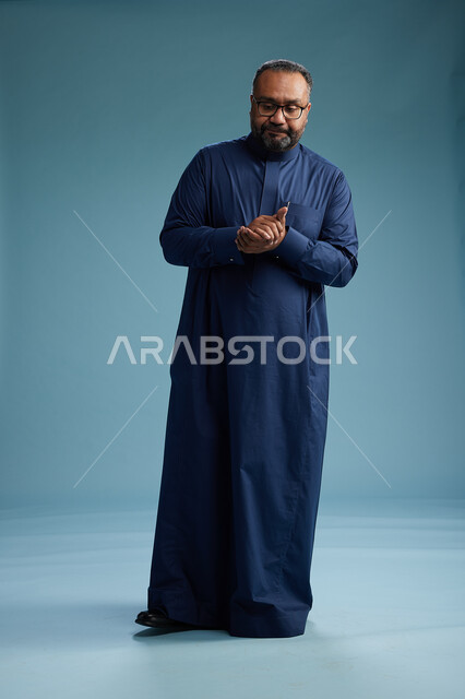 بورتريه لرجل سعودي يقف مرتدياً ثوب كحلي يقوم بايماءات وحركات متنوعه, الوقوف بكامل الجسم, خلفيه زرقاء