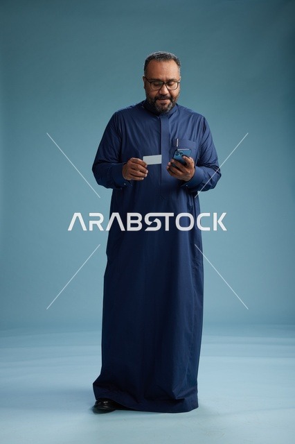 بورتريه لرجل سعودي يرتدي ثوباً أزرق ينظر إلى بطاقة عمل ممسكاً بالهاتف المحمول, الجسم بالطول الكامل, خلفيه زرقاء