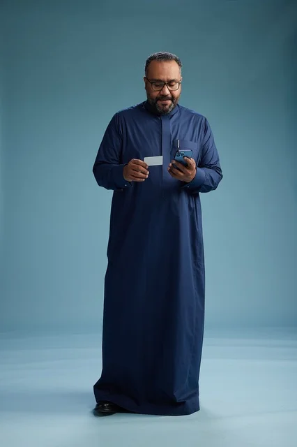 A portrait of a Saudi man wearing a blue thobe looking at a business card while holding a mobile phone, full body, blue background.