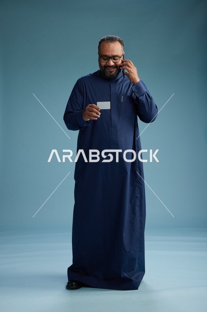 بورتريه لرجل سعودي يرتدي ثوباً أزرق ينظر إلى بطاقة عمل ممسكاً بالهاتف المحمول, الجسم بالطول الكامل, خلفيه زرقاء