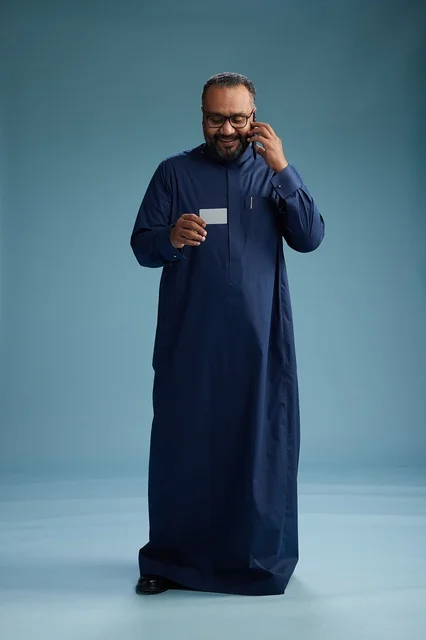 A portrait of a Saudi man wearing a blue thobe looking at a business card while holding a mobile phone, full body, blue background.
