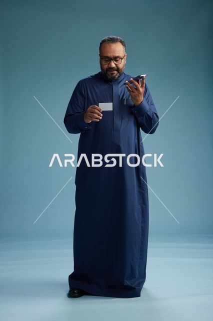 بورتريه لرجل سعودي يرتدي ثوباً أزرق ينظر إلى بطاقة عمل ممسكاً بالهاتف المحمول, الجسم بالطول الكامل, خلفيه زرقاء