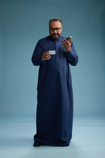 A portrait of a Saudi man wearing a blue thobe looking at a business card while holding a mobile phone, full body, blue background.