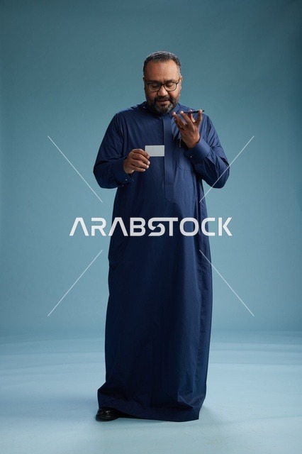 بورتريه لرجل سعودي يرتدي ثوباً أزرق ينظر إلى بطاقة عمل ممسكاً بالهاتف المحمول, الجسم بالطول الكامل, خلفيه زرقاء